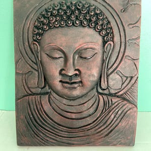 May include: A rectangular, reddish-brown relief sculpture of Buddha with closed eyes. The sculpture features detailed carving of the Buddha's face, hair, and robes, with a halo and floral accents. The artwork is likely made of a durable material.