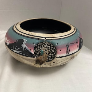 May include: A hand-painted, oval-shaped bowl with a black interior and a cream-colored exterior. The rim features a teal and pink landscape design with a dreamcatcher and feather attached. The bowl is likely used for decorative purposes.