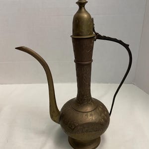 May include: An antique brass pitcher with a long spout and a decorative handle. The pitcher features intricate floral patterns etched into its surface. The design includes a tall, pointed lid and a rounded base, showcasing a vintage aesthetic.
