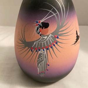 May include: A hand-painted, egg-shaped vase with a Native American design. The vase features a gradient background of peach, purple, and black. The design depicts a figure with outstretched arms, adorned with gray, red, and blue details.