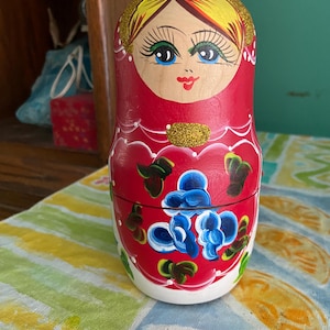 May include: A red Matryoshka doll with a painted face, yellow hair, and blue eyes. The doll has a gold glitter accent and is decorated with blue and green floral designs. The doll is sitting on a colorful striped tablecloth.