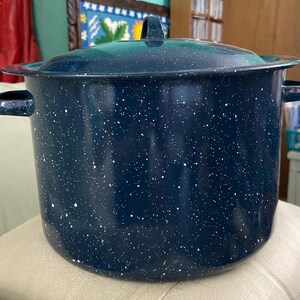 May include: A large, navy blue speckled enamelware pot with a matching lid. The pot has two side handles and a handle on the lid. The pot is sitting on a beige surface.