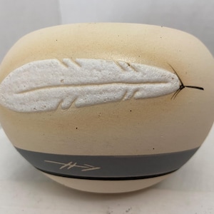 May include: Cream-colored ceramic bowl with a white feather design and a gray band. The feather is textured and raised, with a black line extending from the tip. A gray band with arrow design encircles the base.