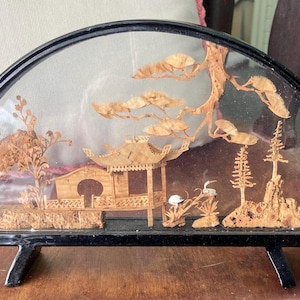 May include: A decorative diorama featuring a detailed miniature landscape scene. The artwork is enclosed in a black, arched frame and depicts a pagoda, trees, and birds, all crafted from light-colored wood.