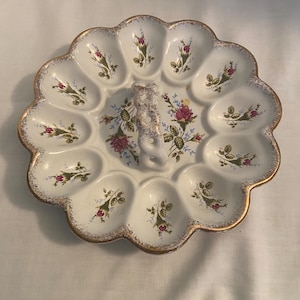 May include: A white ceramic egg serving platter with a scalloped edge and gold trim. The platter features twelve oval indentations, each decorated with a small pink rosebud motif. A central twisted handle with gold accents sits in the middle.