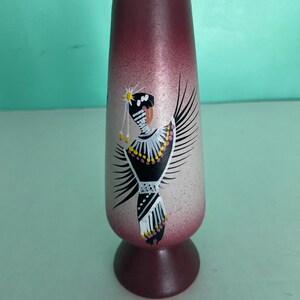 May include: A tall, tapered vase with a pedestal base, featuring a gradient finish that transitions from deep maroon at the top and bottom to a lighter, speckled beige in the center. A painted figure in black, white, and yellow adorns the front.