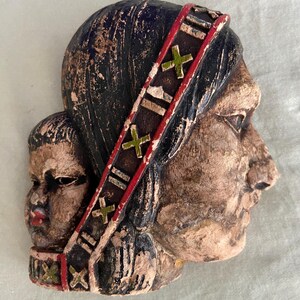 May include: A vintage, weathered sculpture depicting a Native American woman and child in profile. The woman wears a headband with red and green accents. The sculpture is primarily brown with black and blue details.