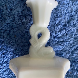 May include: A white, vintage-style candle holder. The candle holder features a dolphin design on the stem and a fluted top. The base is rectangular. The candle holder is made of a milky white glass.
