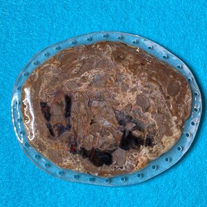 May include: Oval-shaped, polished stone with a clear resin border. The stone features a mix of brown, gray, and black patterns, resembling a cross-section of a tree trunk. The resin border has small, evenly spaced holes.