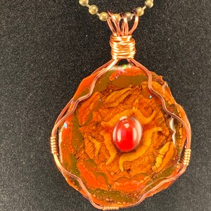 May include: A handcrafted pendant with a unique, organic design. The pendant features a central red stone, surrounded by a textured, earthy-toned material, encased in copper wire. It hangs from a delicate, beaded chain, creating a striking piece of jewelry.