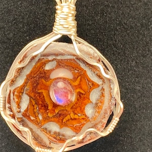 May include: A round pendant featuring a brown and white patterned stone with a central iridescent cabochon, encased in a silver-colored wire-wrapped frame. The pendant hangs from a thin, twisted silver-colored metal chain necklace.