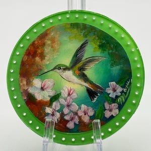 May include: A round, green-edged decorative glass piece featuring a painted hummingbird in flight. The artwork includes white and pink flowers, set against a backdrop of green and brown foliage. The piece is displayed on a clear stand.