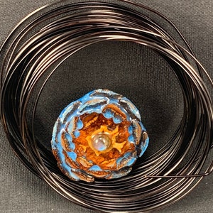 May include: A handcrafted, floral-shaped pendant with blue and brown enamel details, centered with a small, iridescent bead. The pendant is encircled by a coil of dark, metallic wire, creating a unique jewelry component.