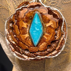 May include: A handcrafted pendant featuring a central, diamond-shaped turquoise stone set within a natural, brown, textured material. The pendant is encased in a silver wire frame, creating a unique, nature-inspired accessory.