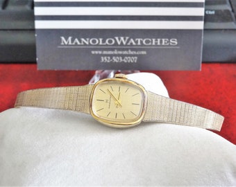 Puede incluir: Un reloj de pulsera dorado con una esfera cuadrada y una pulsera de metal plateado. La esfera del reloj tiene marcadores de hora en números romanos negros y agujas negras. La pulsera del reloj tiene un patrón texturizado.