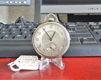 May include: A silver pocket watch with an Elgin face and black hands. The watch is on a clear stand and has a tag that reads "Elgin 1927, 15 Jewels, Size 12, G.F. Case".