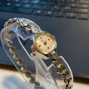 May include: A silver wristwatch with a white face and red accents. The watch has a silver band with a unique design.