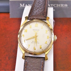 May include: A gold-toned wristwatch with a white face and black leather strap. The watch face has black Roman numeral hour markers and black hands. The watch face also has the text "Bennus 25 Jewels Self-Winding".