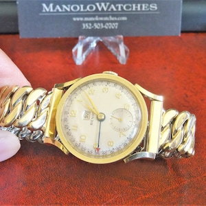 May include: A gold wristwatch with a white face and a gold chain link band. The watch face has black Roman numeral hour markers, a date window, and a small seconds dial. The watch is on a red surface.