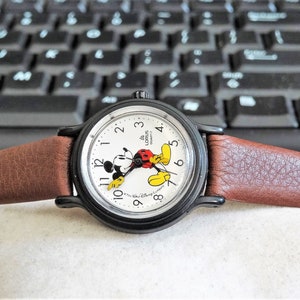 May include: A brown leather wristwatch with a black face and Mickey Mouse design. The watch face has white numbers and the words "Lorus Disney" printed on it.