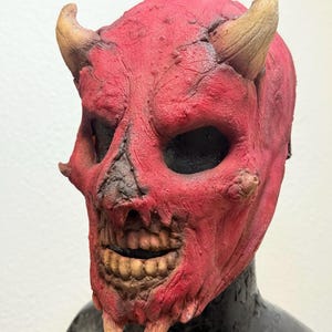 Op de afbeelding: Een rood duivelsmasker met bruine hoorns en een skeletachtig gezicht. Het masker heeft grote oogkassen, een brede grijns met zichtbare tanden en een getextureerd oppervlak. Het masker is ontworpen om gedragen te worden.