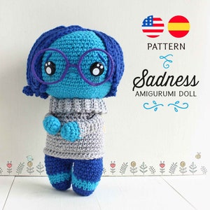 May include: Crocheted amigurumi doll of the character Sadness from the movie Inside Out. The doll is blue with a gray sweater and purple glasses. The doll is standing on a white wooden surface.