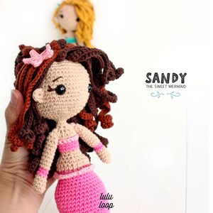Peut inclure: Poup&eacute;e sir&egrave;ne en crochet avec des cheveux bruns, une queue rose et un haut rose. La poup&eacute;e tient une fleur rose dans ses cheveux. La poup&eacute;e est &eacute;tiquet&eacute;e "Sandy The Sweet Mermaid".