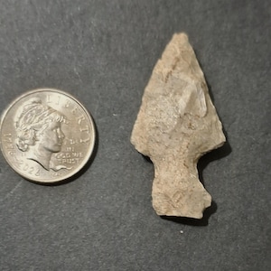 May include: A light tan, chipped stone arrowhead with a triangular blade and a notched stem base, positioned next to a silver-colored United States quarter coin for scale, showing the coin's profile of a person and the text Liberty and 1776-2026.
