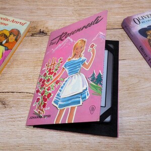Könnte beinhalten: Eine rosa E-Reader-Hülle mit dem Buchcover von Das Rosenresli von Johanna Spyri, das eine Person in einem blau gestreiften Kleid mit weißer Schürze neben roten Rosen zeigt, flankiert von den Büchern Der zweite Anruf für Simone und Oliver Twist.