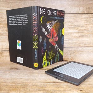 Könnte beinhalten: Ein deutschsprachiges Buch mit dem Titel "DIE KLEINE HEXE" mit einer Hexenillustration, neben einem schwarzen E-Reader. Das Buch hat einen schwarzen Einband mit weißem Text und einer farbenfrohen Illustration.
