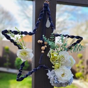 May include: A navy blue macrame star with white and light blue flowers, greenery, and wooden beads hanging from it.