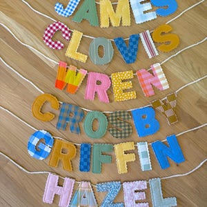 May include: A selection of fabric name banners spelling out "JAMES", "CLOVIS", "WREN", "CROSBY", "GRIFFIN", and "HAZEL". Each letter is made from a different patterned fabric, including gingham and floral prints. The banners are strung on white cords.