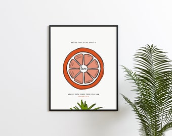 May include: A white framed print with a red and pink grapefruit illustration. The grapefruit is divided into sections with words like "love", "joy", "peace", and "kindness" written inside each section. The text "BUT THE FRUIT OF THE SPIRIT IS" is at the top of the print and the text "AGAINST SUCH THINGS THERE IS NO LAW" is at the bottom.