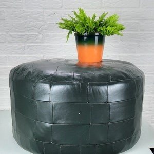 Op de afbeelding: Een donkergroene leren poef met een vierkant gestikt patroon. Een kleine plant in een pot met groene bladeren staat op de poef.