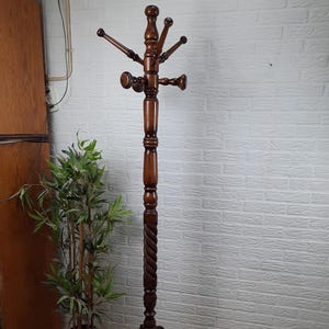 Op de afbeelding: Een donkerbruine houten kapstok met een gedraaide voet en vijf haken. De kapstok staat op een betegelde vloer voor een witte bakstenen muur.