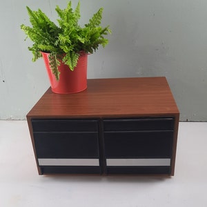 Könnte beinhalten: Ein Holzschrank mit zwei schwarzen Türen und silbernen Zierleisten. Ein kleiner roter Topf mit einer grünen Pflanze steht oben drauf. Der Schrank hat eine warme braune Oberfläche.