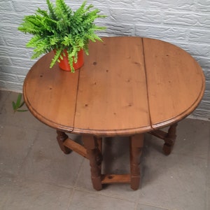 May include: A round, wooden drop-leaf table with a warm brown finish. The table has four legs and a hinged top that can be folded down. A potted green plant sits on the table. The table is approximately 76 cm in diameter.