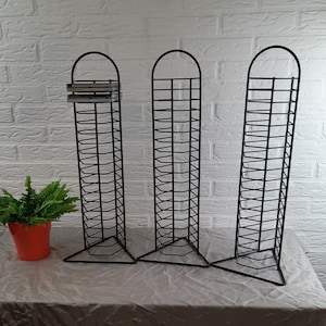 Puede incluir: Tres torres de almacenamiento de CD de metal negro sobre un mantel blanco. Cada torre tiene varios estantes para guardar CD.