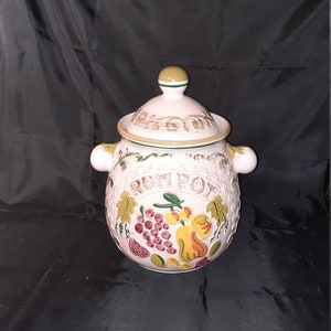 May include: A ceramic jar with a lid, featuring a light pink base and a design of fruits and the word "POMPON" in a banner. The jar has two small handles and a green and yellow trim. The lid has a small green and yellow knob.