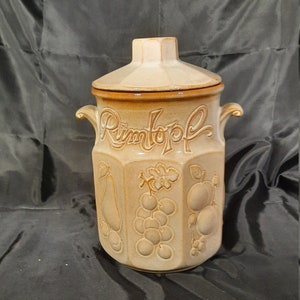 May include: A brown ceramic jar with a lid, featuring the word "Rimtopf" in cursive script. The jar is decorated with embossed fruit designs, including pears, grapes, and apples.