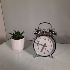 以下が含まれることがあります： 白い文字盤と黒い数字の銀色の目覚まし時計。時計の上部にベルが2つあり、7時5分に設定されています。時計は薄い緑色の表面の上に置かれています。