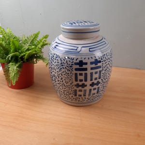 May include: A ceramic ginger jar with a lid, featuring a blue and white floral and geometric design. The jar has a rounded shape and is placed on a wooden surface. The design includes a double happiness symbol.
