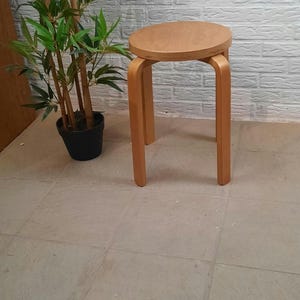 Può includere: Uno sgabello in legno chiaro con seduta rotonda e quattro gambe curve. Lo sgabello è posizionato davanti a un muro di mattoni bianchi. Una pianta di bambù in vaso è a sinistra dello sgabello.