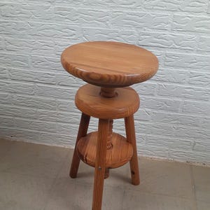 May include: A wooden adjustable stool with a round seat and a lower circular support. The stool has three legs and a central threaded column for height adjustment. The wood has a natural finish.