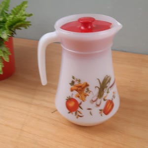 Op de afbeelding: Witte melkglazen kan met een rode deksel en handvat. De kan is versierd met kleurrijke illustraties van groenten. Een vintage keukenartikel.