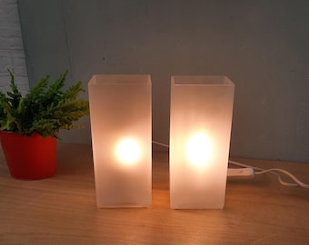 May include: Two frosted glass lamps with a warm glow, rectangular in shape. The lamps are illuminated, casting a soft light. A small potted plant in a red pot is visible on the left. The lamps are on a wooden surface.