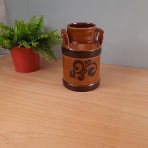 Op de afbeelding: Een bruine keramische melkkan met een bloemenmotief. De kan heeft twee handvatten en een bruine glazuur.