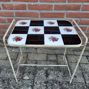 May include: A white metal side table with a black and white checkered top. The top features a floral design with red, white, and green flowers. The table has a single shelf.