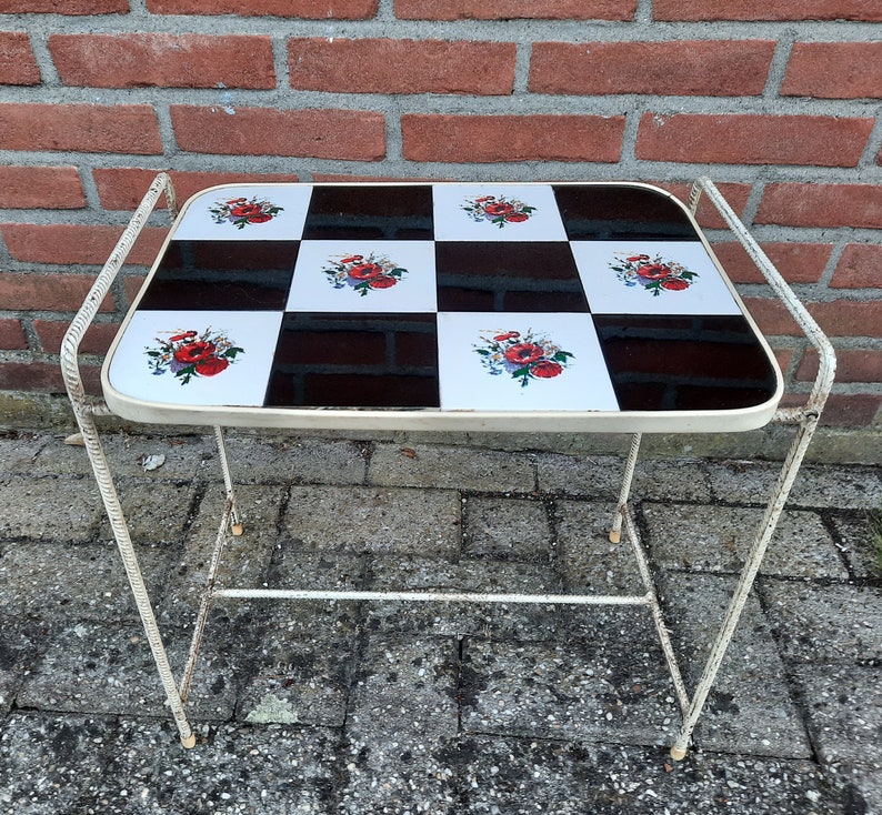 May include: A white metal side table with a black and white checkered top. The top features a floral design with red, white, and green flowers. The table has a single shelf.