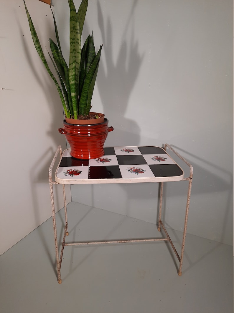 May include: A white metal side table with a black and white checkered top. The table has a red ceramic planter with a green snake plant on top.